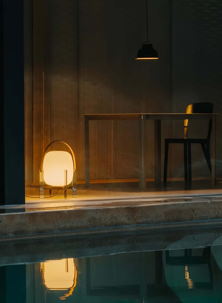 Modern interior with a table and chair by a pool, illuminated by a Alix Cesta Metálica Basket Lamp with chrome legs and frosted glass