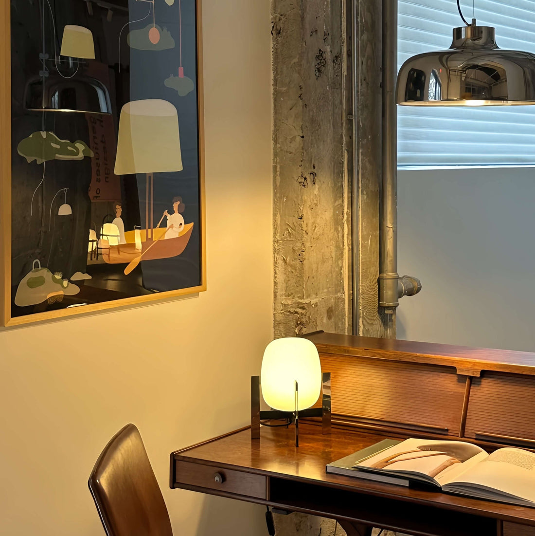 Wooden desk withAlix Cesta Metálica Basket Lamp with chrome legs and frosted glass and open book in a room with a painting on the wall.