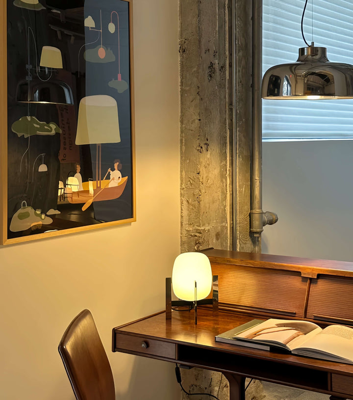 Wooden desk withAlix Cesta Metálica Basket Lamp with chrome legs and frosted glass and open book in a room with a painting on the wall.