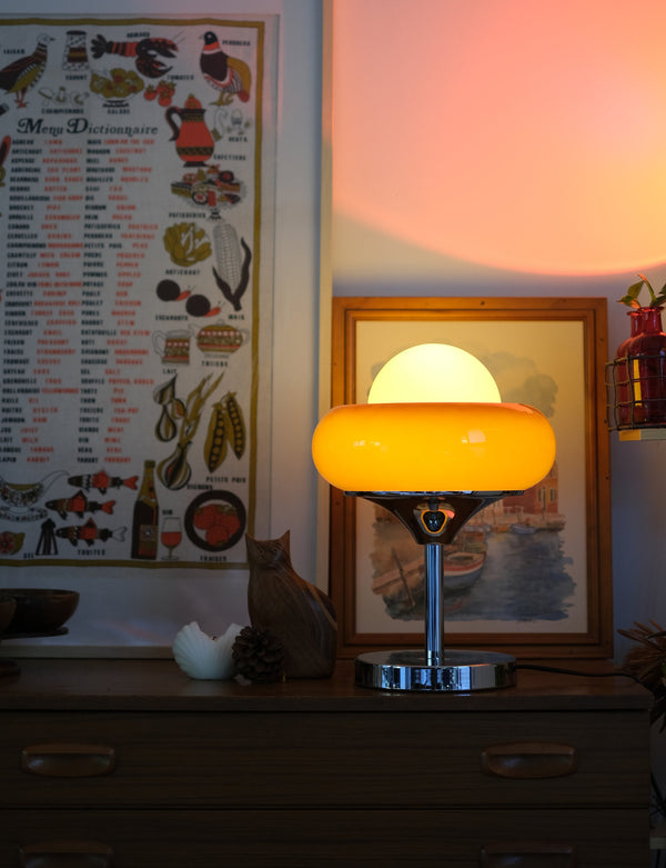 Egg Tart Table Lamp with orange dome glass shade and white inner globe with a warm glow on a desk next to a cat and decorative items.