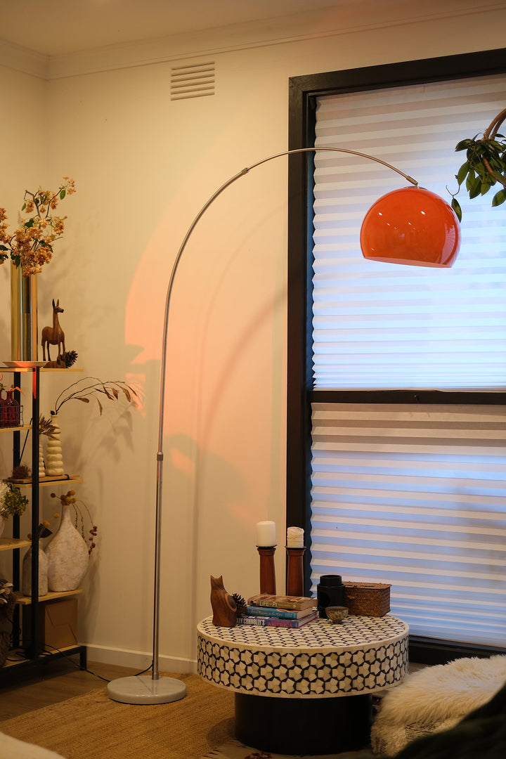 Sneedville+ Bauhaus Arc Floor Lamp with chrome arc and marble base in white shade Orange lamp casting a warm glow in a dimly lit room with posters and decor. Living room with a tall floor lamp, small table, and window blinds.