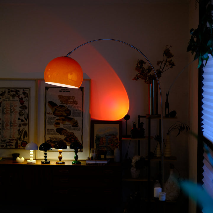 Sneedville+ Bauhaus Arc Floor Lamp with chrome arc and marble base in white shade Orange lamp casting a warm glow in a dimly lit room with posters and decor.