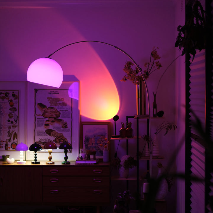 Sneedville+ Bauhaus Arc Floor Lamp with chrome arc and marble base in white shade Orange lamp casting a warm glow in a dimly lit room with posters and decor. Room interior with a glowing lamp casting a pinkish-purple light.