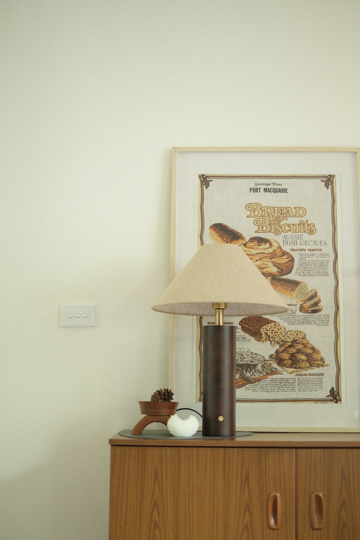 Morgan Table Lamp in Walnut with linen shade on a wooden cabinet with a framed poster of bread in the background.